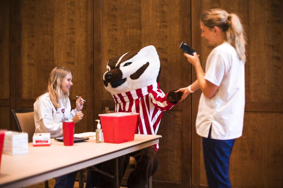 Bucky Badger is seated at a table receiving a flu vaccine from a UW Nursing student wearing a white coat.