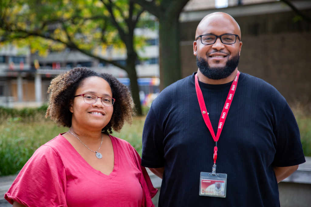 Josie Montañez-Tyler and Corey Steele, both UHS providers, are pictured outside with a tree in the background. Josie wears a red shirt and Corey wears a black shirt with a red UHS lanyard. 