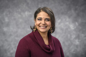 Dr. Josyula is pictured in a purple shirt with gold and purple earrings in front of a gray background.