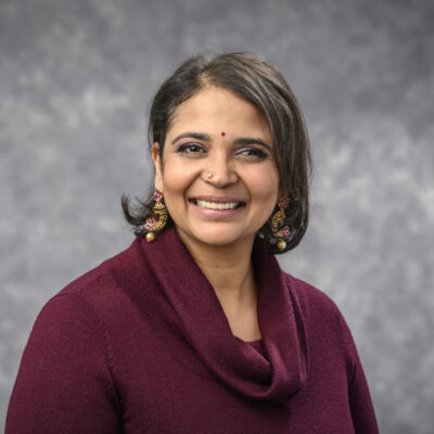 Dr. Josyula is pictured in a purple shirt with gold and purple earrings in front of a gray background.