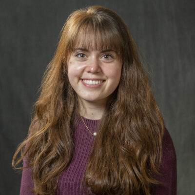 Grayson Murray has long brown hair and is wearing a maroon shirt and silver necklace.