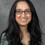 Dr. Nasia Safdar has long brown hair, wears glasses, and is wearing a blue and green shirt.