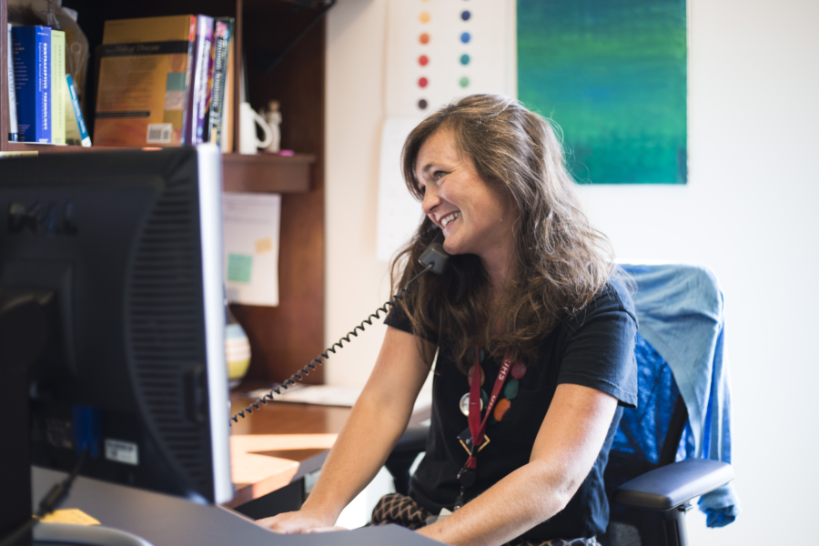 A nurse with long brown hair smiles as she takes a telehealth phone call at her computer.