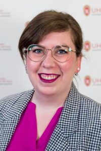 Katrina Welborn is a woman with short brown hair and glasses. She wears magenta lipstick, a magenta blouse, and a checkered blazer.