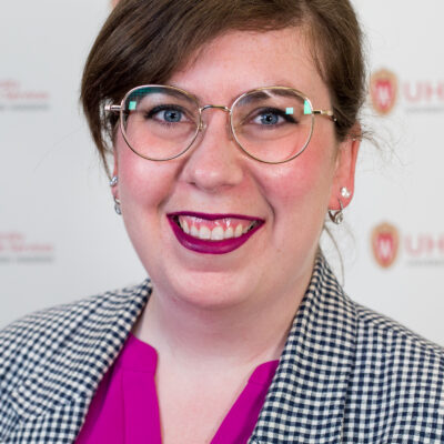 Katrina Welborn is a woman with short brown hair and glasses. She wears magenta lipstick, a magenta blouse, and a checkered blazer.