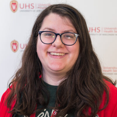 Andie Schwabe has long brown hair, wears glasses, and is pictured in a red sweater and green t-shirt.