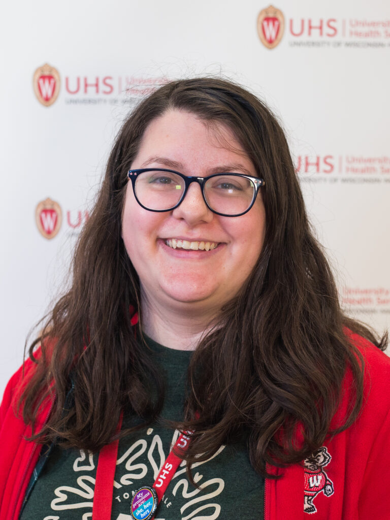 Andie Schwabe has long brown hair, wears glasses, and is pictured in a red sweater and green t-shirt.