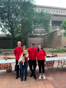 Photo of Badger Recovery students standing in front of tables for tailgate event with a Corgi