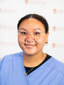 A woman with brown hair and thin-rimmed glasses wears a blue scrub top over a black shirt.