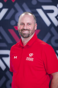 A man with a short beard is wearing a red shirt and standing in front of a background with the RecWell logo on it.