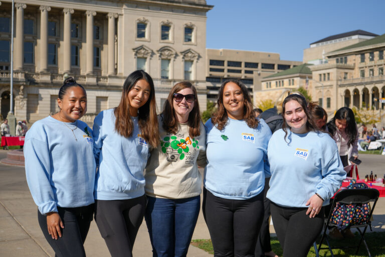 Five people stand on Library Mall with their arms around each other smiling to the camera.