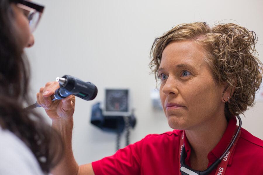 A nurse wearing a red shirt uses a medical instrument to look in a student's mouth.