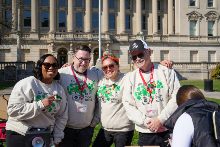 Four mental health providers in matching beige sweatshirts stand smiling together in front on Library Mall