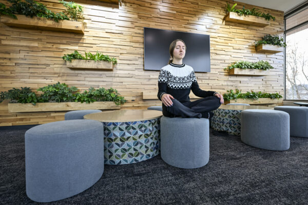 A person sits cross-legged, meditating on a round, gray ottoman in a modern room with wood paneling and green plants, conveying a serene atmosphere.