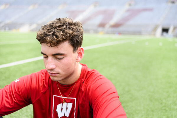 Young man in a red shirt sits on a football field. The background is blurred stadium seats