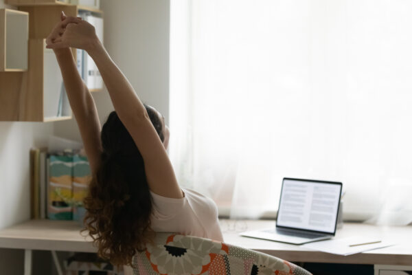 A person stretches at a desk by a window, arms raised, in a bright home office. A laptop displays text, and shelves with books are visible, conveying a relaxed mood.