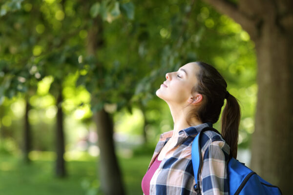 A woman with a ponytail and plaid shirt stands peacefully with closed eyes in a sunlit forest, wearing a blue backpack, enjoying nature.