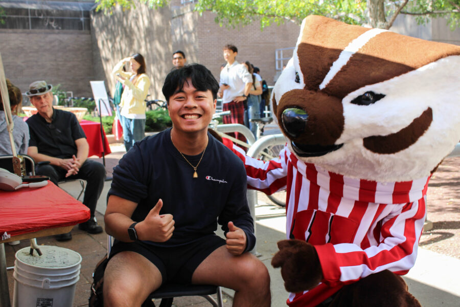 A student smiles and gives a thumbs up after receiving their flu vaccine. The student is accompanied by Bucky Badger.