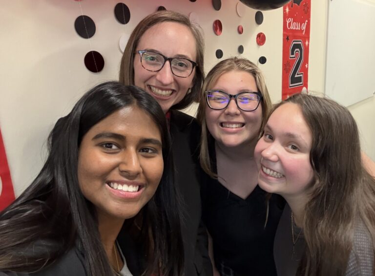 Four people stand together smiling in front of a graduation balloon display
