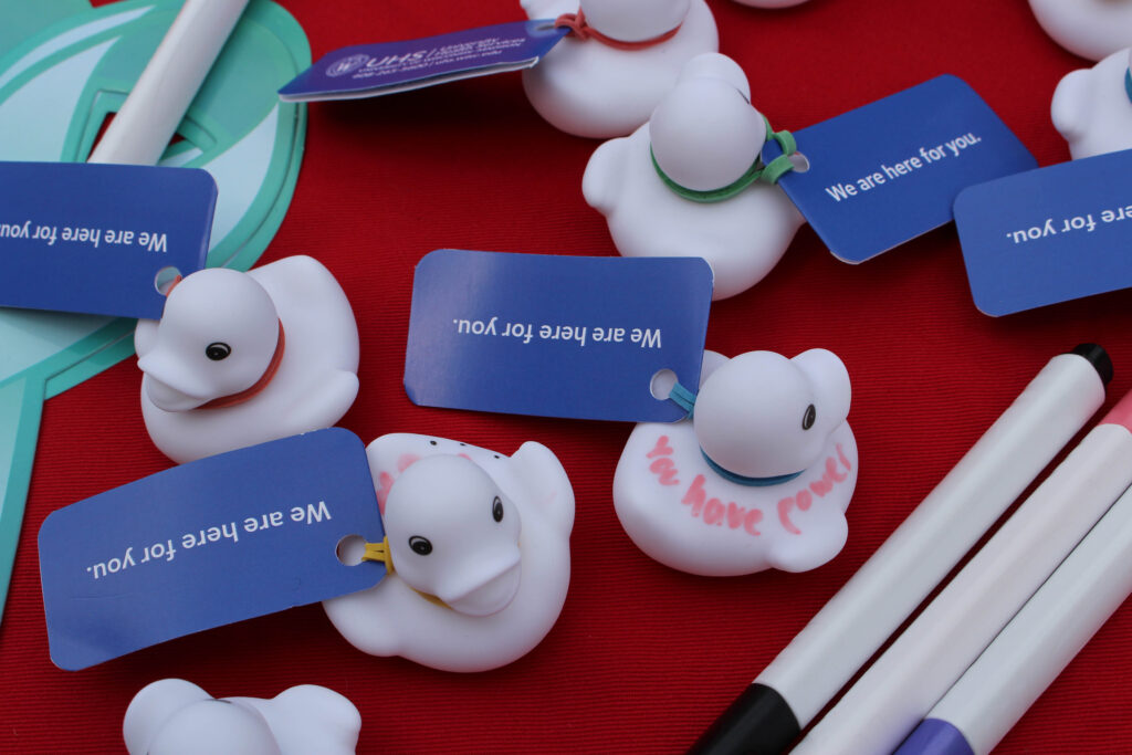 White rubber ducks on a table with a tag attached that says "we are here for you". One duck is decorated with marker that says "you have power"
