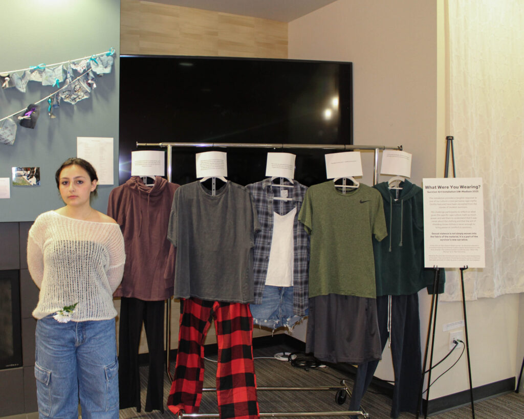 Sofia More posing with their art exhibit titled "What were you wearing." Clothing is hung on a rack next to Sofia.