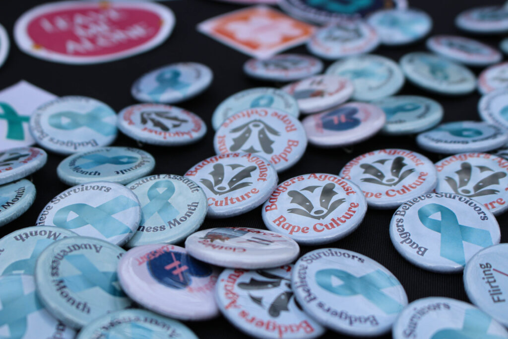 Button pins on a black table with different designs that say "Believe Survivors", "Sexual Assault Awareness Month", and "Badgers Against Rape Culture"