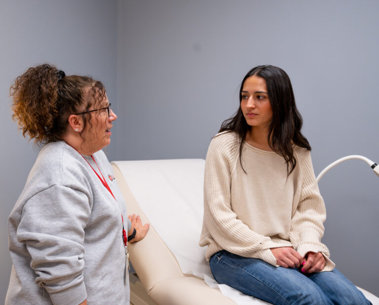 Survivor Services Advocate Lea Ann Harris converses with student who is seated upright with their hands in their lap on the medical table