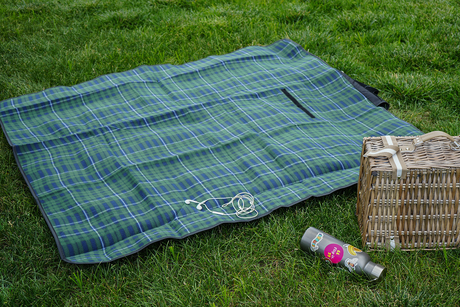 Green plaid picnic blanket on grass with wired headphones, water bottle, and pinic basket.