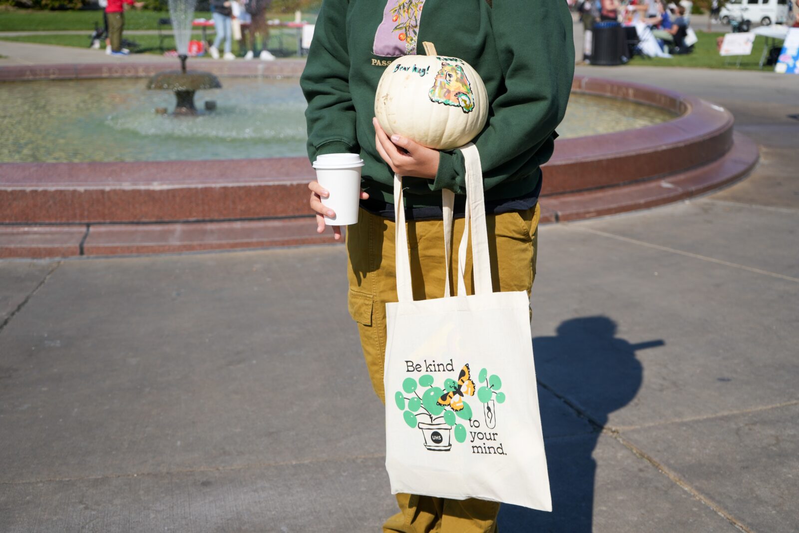 Student holds the giveaway tote bag, painted pumpkin, and a coffee that the mental wellbeing resource fair.