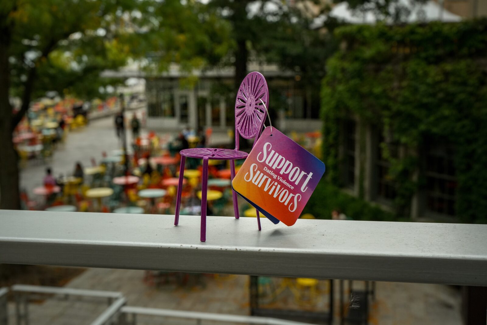 The 2026 mini terrace chair sitting on top of a railing at the Memorial Union Terrace with terrace table and chairs in the background.